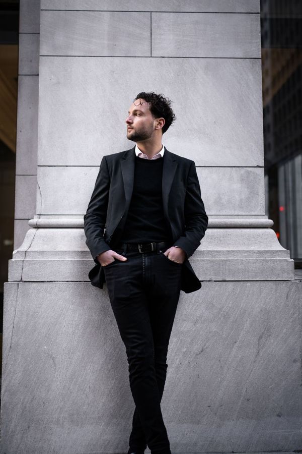 Man in a blazer leaning against stone with hands in his trouser pockets